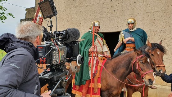 Drei Pferde mit Reitern in römischer Legionärskleidung. Davor eine große Videokamera mit Kameramann und Assistent. Im Hintergrund die Sandsteinwand eines Gebäudes