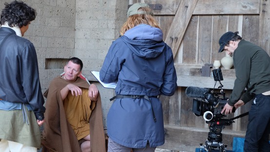 In einer Art Schuppen mit großem Holztor: Zwei Männer in römischer Kleidung sprechen miteinander. Eine Frau mit Notizbuch schaut zu und rechts steht ein Kamerateam. 