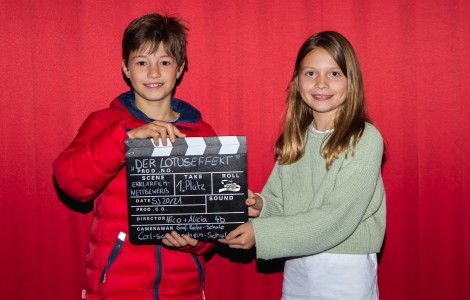 Zwei Kinder stehen vor einer roten Wand im Kino. Sie lächeln und halten eine Filmklappe in der Hand.