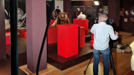 Eine Person in Jeans und Hemd bedient eine große Filmkamera. Vor seiner Kamera stehen ein ausgestopfter brauner Bär und ein weißer Schwan auf roten Emporen, sowie weitere Ausstellungsstücke. 