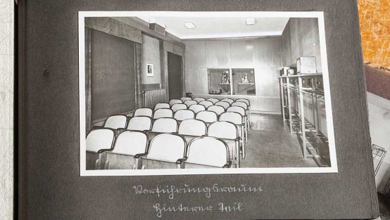 Schwarz-weiss-Foto des Filmvorführraums mit Filmprojektoren, Stuhlreihen in der Mitte und Garderoben an der rechten Wand. Links die abgedunkelten Fenster