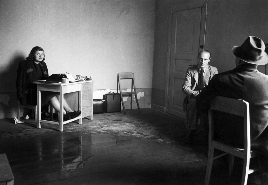 Eine Frau sitzt an einem Schreibtisch und zwei Männer sitzen auf Stühlen, der Boden des Zimmers ist nass