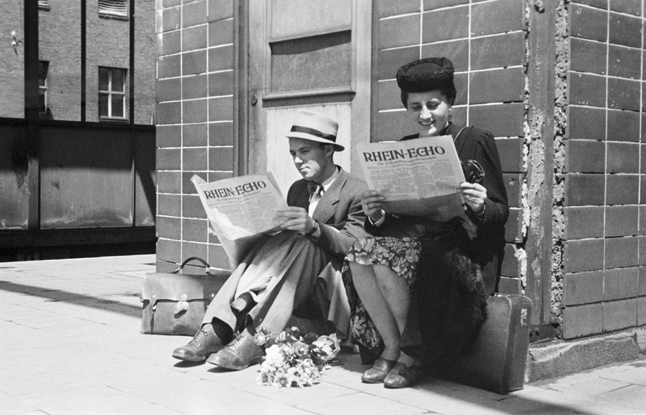 Zwei Menschen sitzen auf dem Bahnsteig und lesen Zeitung