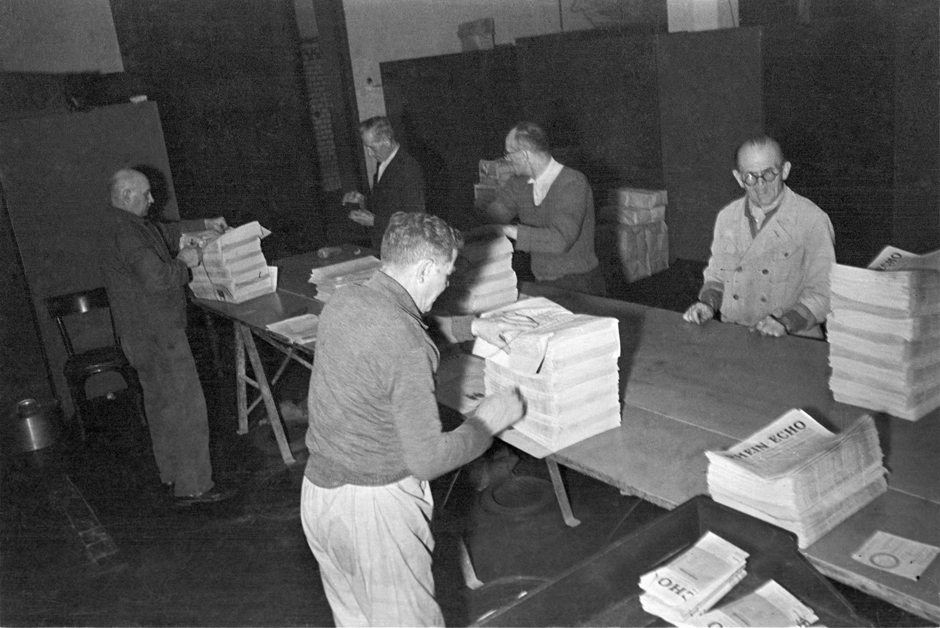  Männer stehen an einem Tisch und bündeln Zeitungen