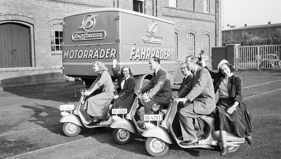 Drei Frauen und drei Männer als Fahrer und Beifahrer auf fabrikneuen Vespa-Rollern