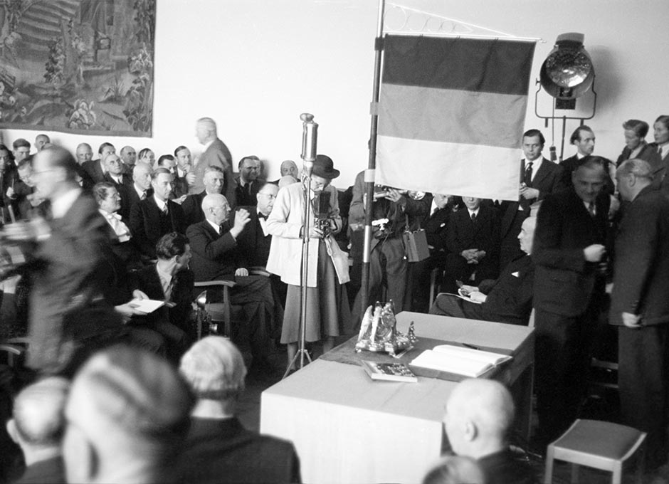 Ein Saal mit Publikum und ein Tisch, neben dem die Flagge der BRD steht.