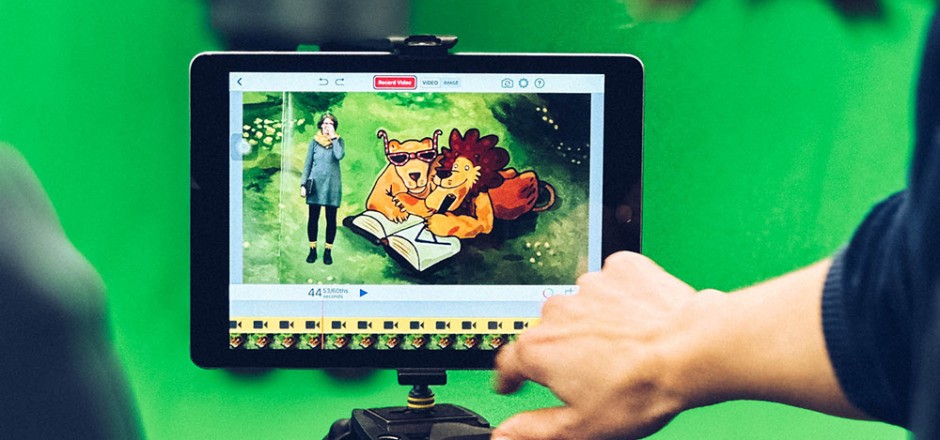 Tablet mit Greenscreen-App: Eine Frau steht neben einem illustrierten Löwenpaar. Der Löwe schreibt in ein Buch.