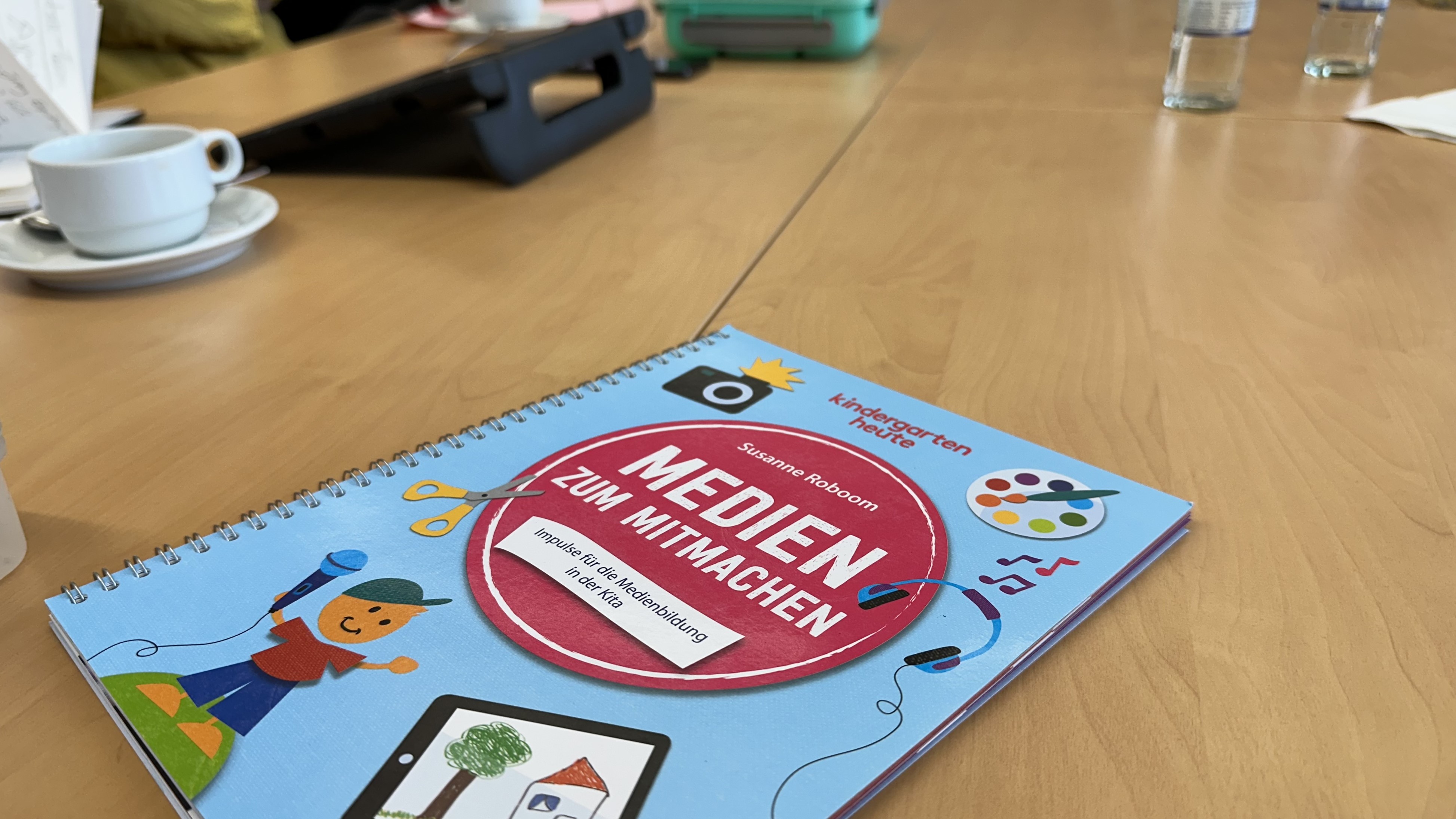  Das Buch "Medien zum Mitmachen" im Vordergrund auf einem belebten Tisch mit Kaffeetassen, Notizbüchern und Wasserflaschen.