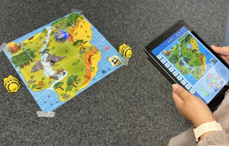  An einem iPad wird ein Bienenroboter gesteuert, der auf einer Land- und Schatzkarte am Boden läuft.