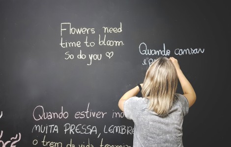 Schülerin steht vor einer Tafel und schreibt Sätze in verschiedenen Sprachen, unter anderem "flowers need time to bloom, so do you".