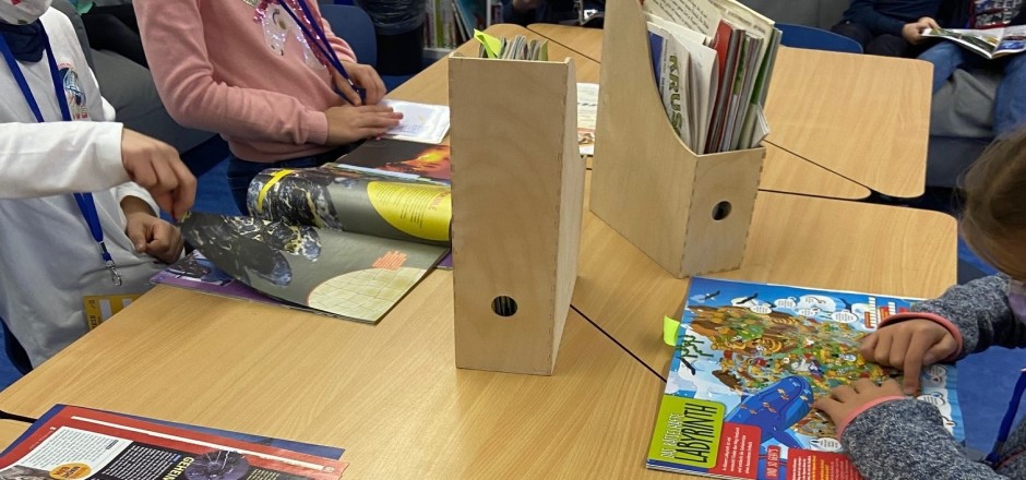 Kinder sitzen an Tischen und auf Sofas und blättern in Zeitschriften. Im Zantrum stehen zwei gefüllte Zeitschriftenhalter aus Holz.