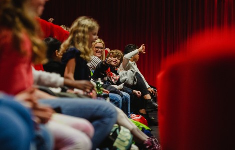 Kinder sitzen in roten Kinosesseln, zeigen den Daumen nach oben und lachen. 