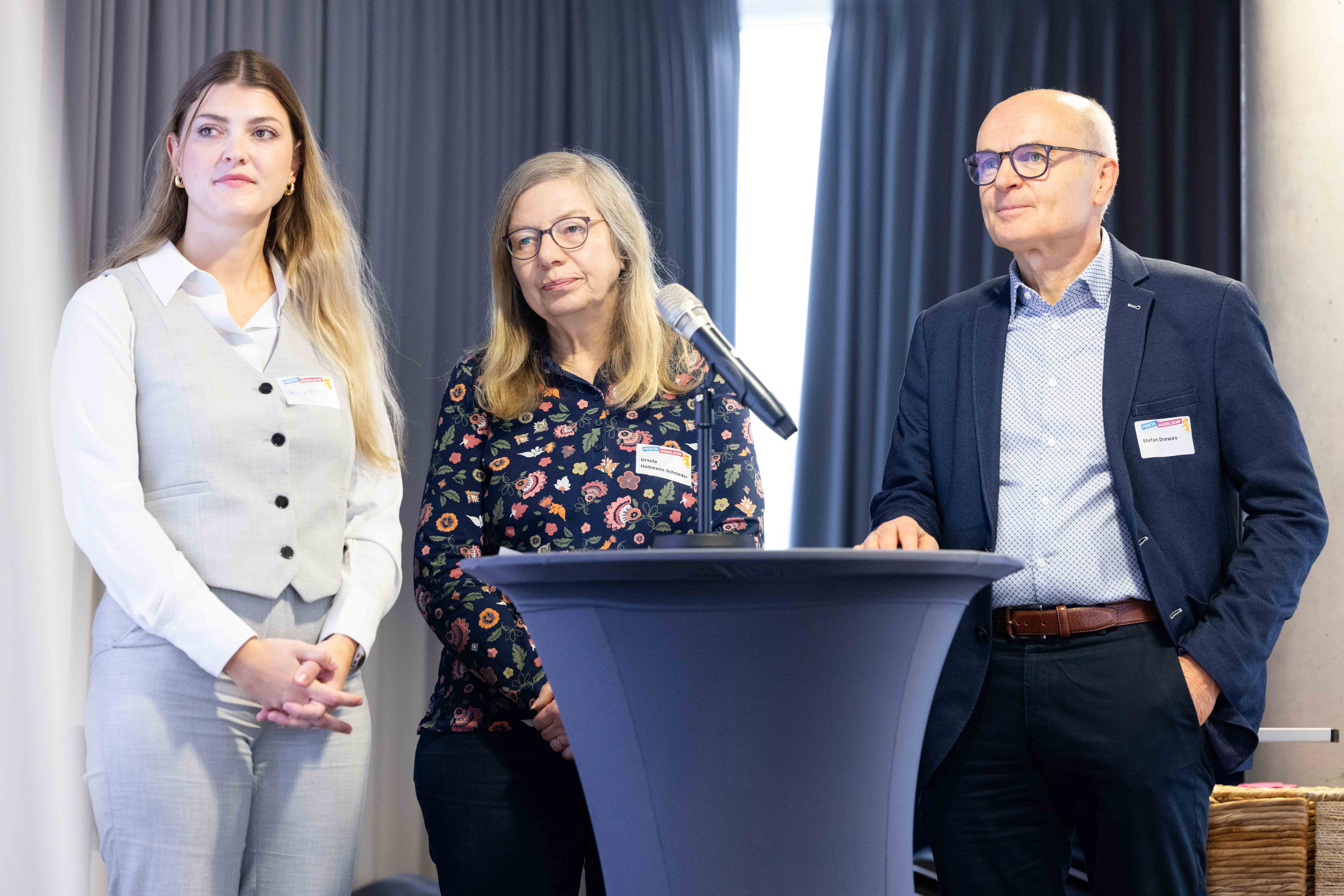 Drei Personen stehen an einem Stehtisch, in der Mitte ein Mikrofon, im Hintergrund Vorhänge.