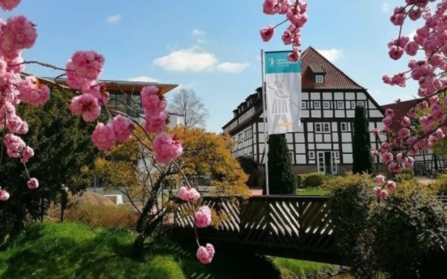 Das Hotel Schnitterhof in Bad Sassendorf mit Blick durch die Äste eines Kirschbaums mit rosanen Kirschblüten auf eine Brücke über einen Bach und das Fachwerkgebäude.