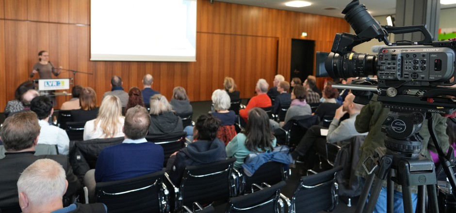 Konferenzraum mit Publikum und einer Sprecherin am Pult.
