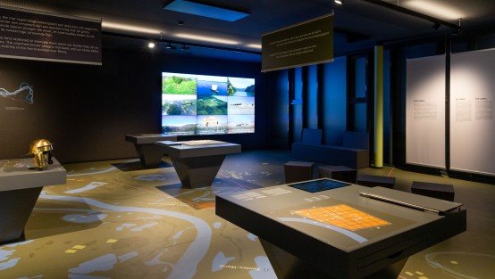 Blick in den Ausstellungsraum mit Infotischen und einer Videowand.