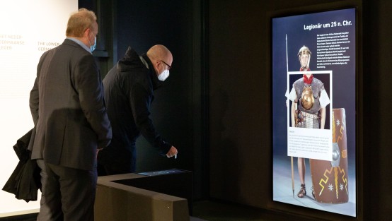 Besucher der Eröffnung der Landesausstellung vor der interaktiven Station mit einem Großbildschirm