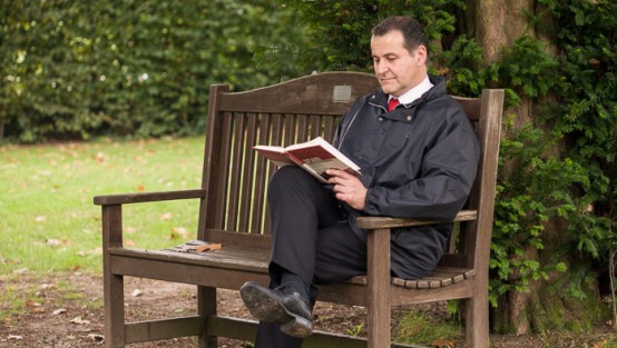 Ein Mann sitzt auf einer Bank und hält ein offenes Buch in der Hand.
