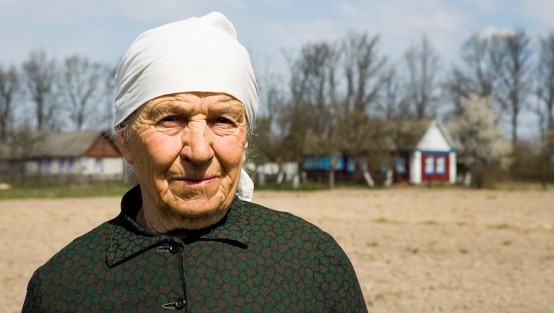 Alte Frau mit weißem Koptuch auf einer Ackerfläche vor kleinen Holzhäusern