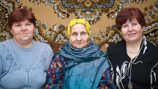 Alte Frau, eingerahmt von ihren beiden Töchtern, vor einem dekorativen Wandteppich