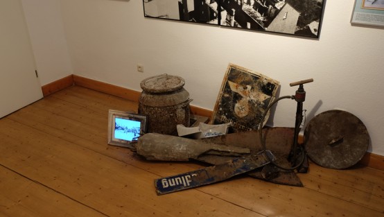 Auf einem Holzboden liegen alte Funde aus dem 2. Weltkrieg, wie z.B. altes Straßenschild oder eine alte Fahrradpumpe. Links vor einem Bottich steht ein digitaler Bilderrahmen, darauf laufen alte historische Bilder.