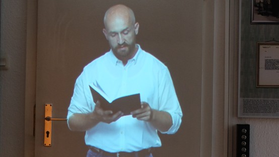 Eine Projektion eines Mann mit kahlem Kopf auf eine weiße Tür. Er hält ein offenes Buch in der Hand.