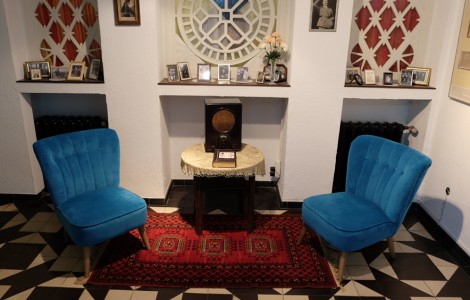 Ein wohnlicher Raum. Zwei blaue Sessel dazwischen auf einem runden Beistelltisch ein Volksempfänger. Auf der Fensterbank im Hintergrund stehen viele alte Fotos.