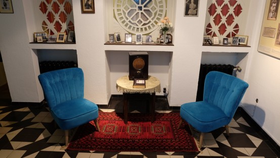 In einem Raum stehen 2 blaue Sessel, in der Mitte ein runder Beistelltisch mit einem Volksempfänger dadrauf.