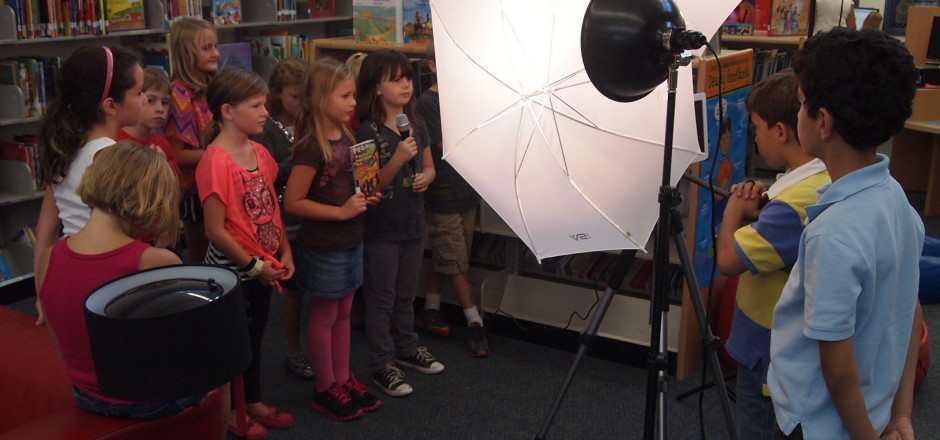 Kinder drehen einen Film in einer Bibliothek. Einige haben ein Mikrofon in der Hand. Vor ihnen stehen große Videoleuchten.