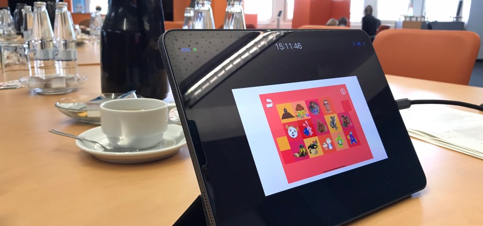 Auf einem Tisch steht ein Tablet. Auf dem Display ist ein Bild aus der Elefanten-App zu sehen: Ein roter Hintergrund mit vielen verschiedenen kleinen Bildern und Symbolen.